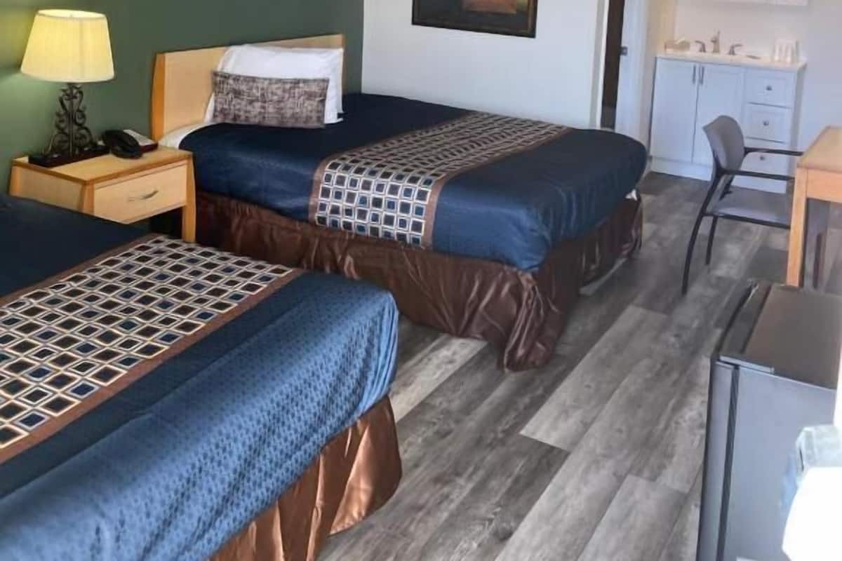 Ein Hotelzimmer mit zwei Betten, einem Nachttisch, einer Lampe, einem gerahmten Bild, einem Spiegel, einem kleinen Schreibtisch und einem Stuhl.
