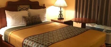 A hotel room with a large bed, a bedside table with a lamp, a framed picture on the wall, and a window with curtains.