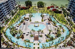 Uma grande piscina circular com piscinas menores e uma fonte central, cercada por espreguiçadeiras e palmeiras.