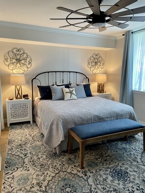 Un dormitorio con cama, mesitas de noche, un banco, ventilador de techo y decoración mural.