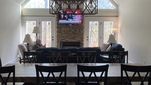 A spacious living room with a stone fireplace, a large TV mounted above it, and a dining area with a glass-top table and chairs. - Serenity, Waterfront Mountain Retreat (Eagle Rock)
