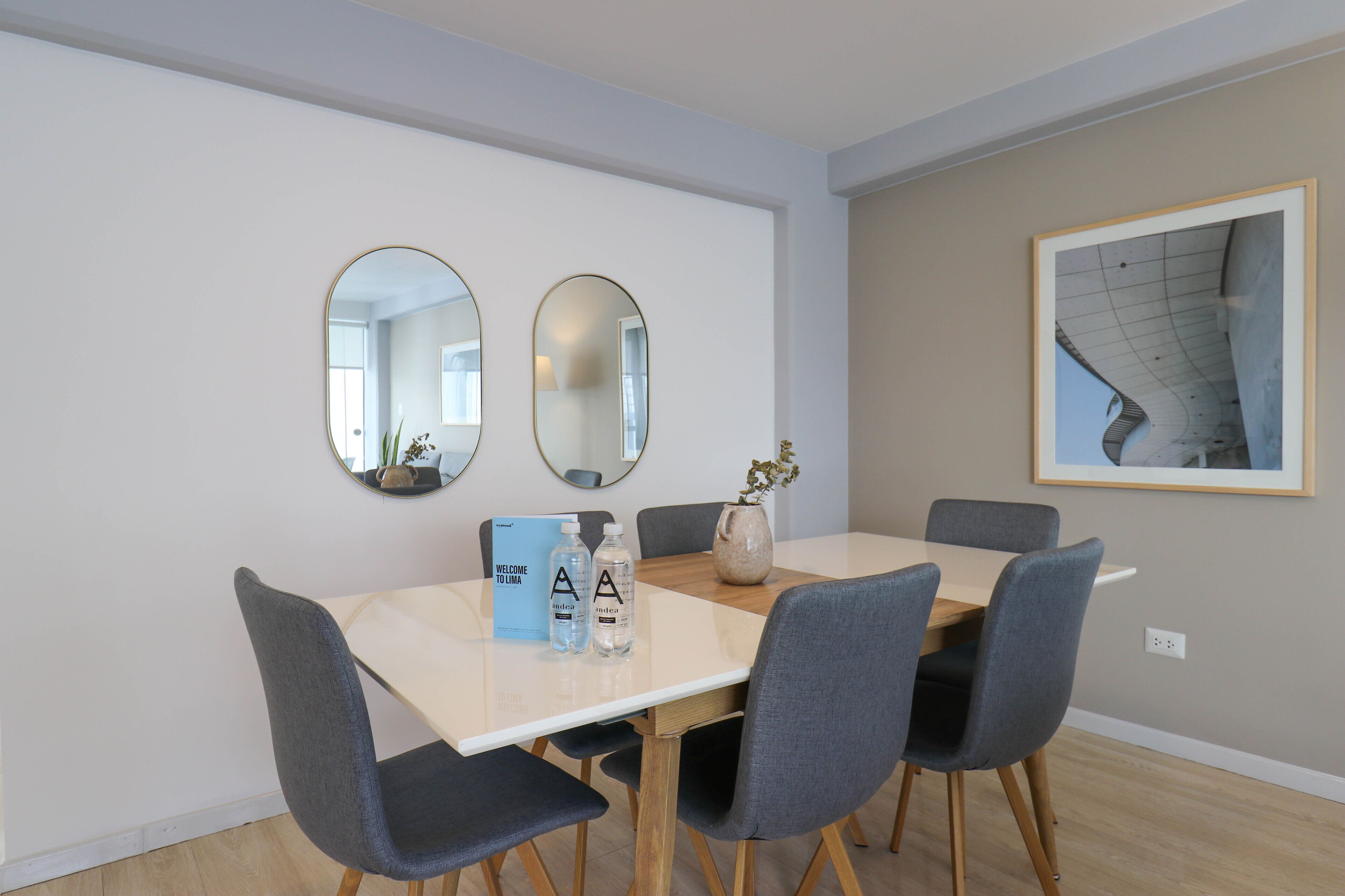 A modern dining area with a white table, four grey chairs, two round mirrors, a framed picture on the wall, and a welcome sign.