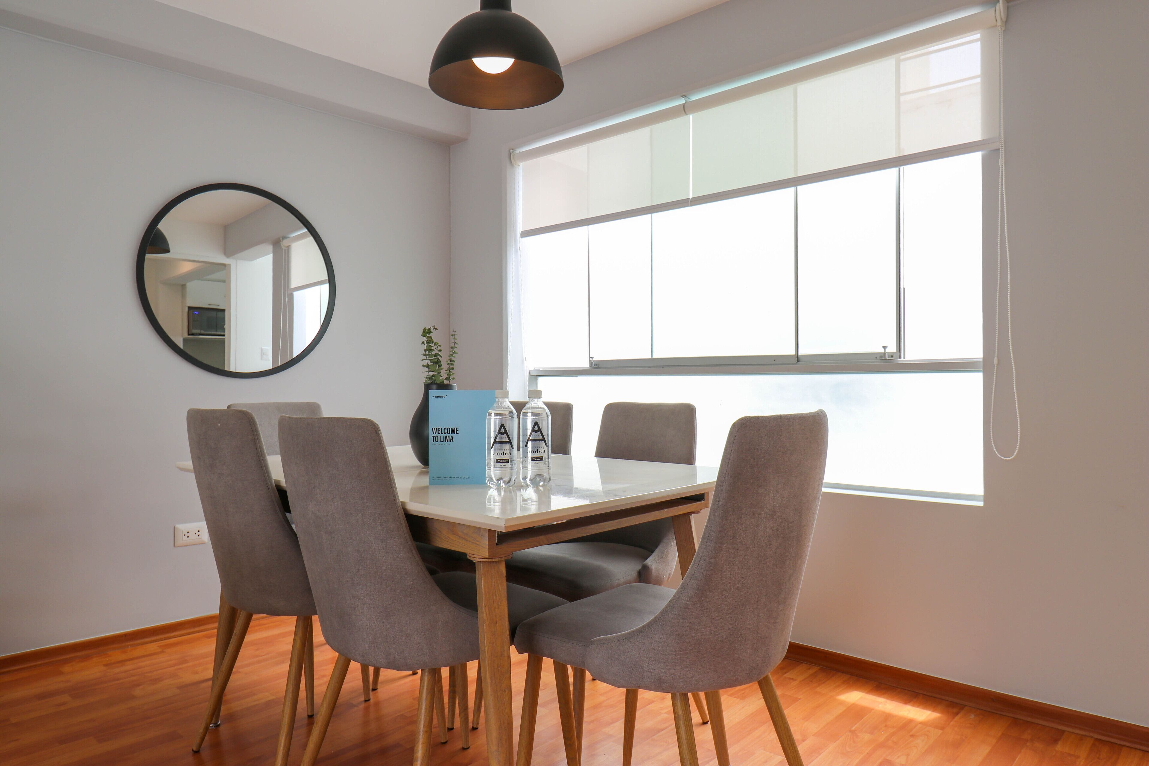 A dining area with a round mirror, a table with four chairs, a blue welcome box, and a large window.