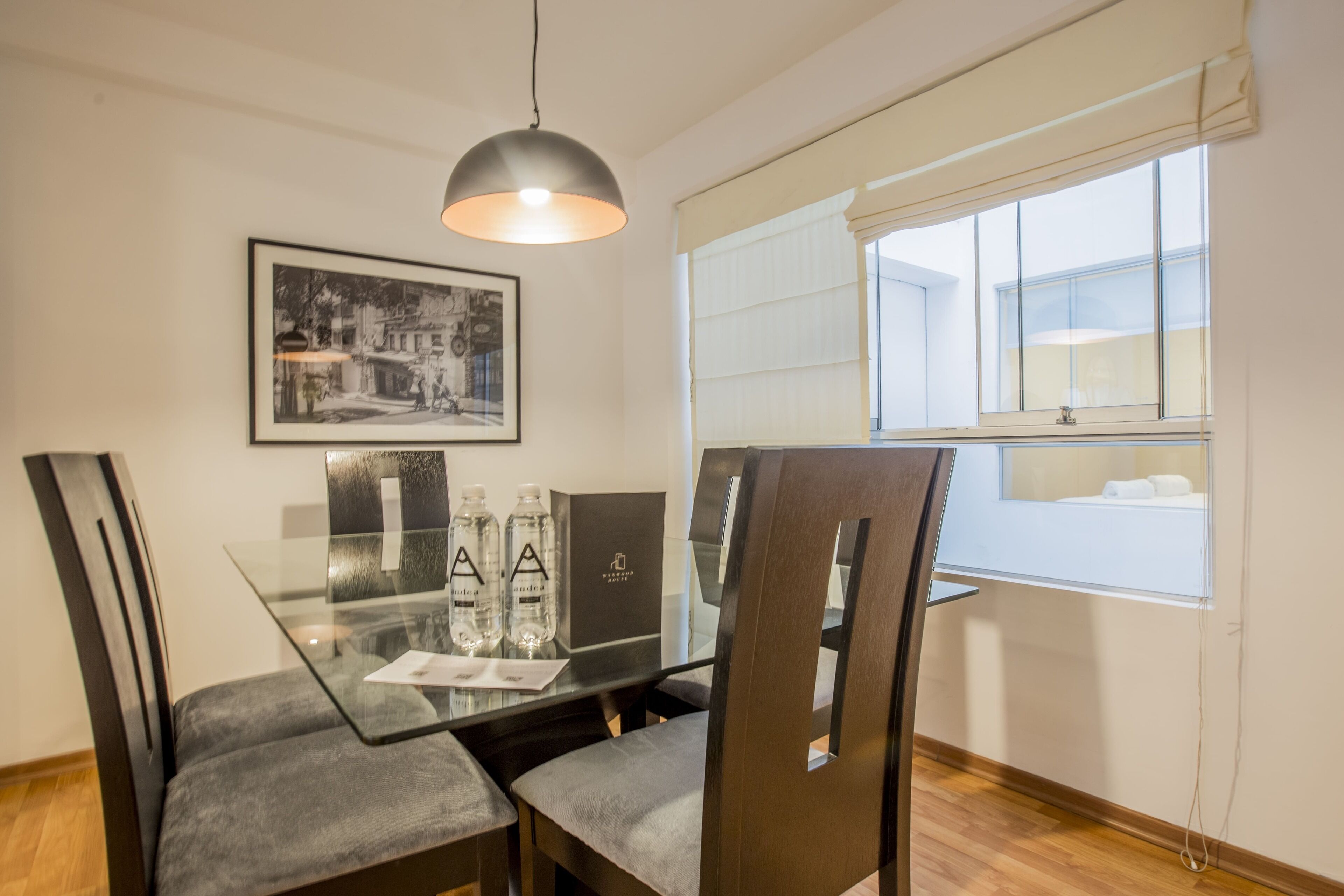 Un coin repas comprenant une table en verre, quatre chaises, un luminaire suspendu, une photographie encadrée accrochée au mur et une fenêtre avec des stores.
