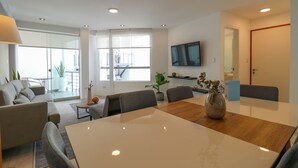 Una sala de estar moderna con comedor, sofá, televisor montado en la pared y un gran ventanal.
