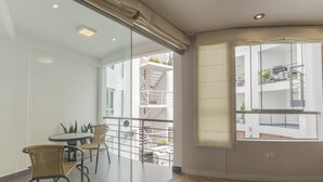 Un balcón moderno con separador de vidrio, una mesa redonda con sillas y vistas a un edificio blanco con balcones.