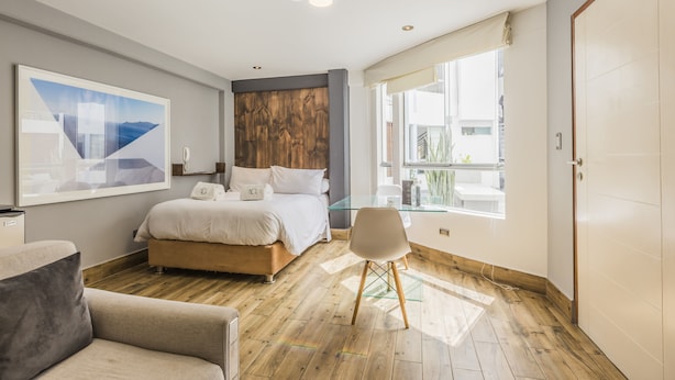 Un dormitorio moderno con cama, escritorio de vidrio y un ventanal grande.