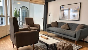 Una sala de estar moderna con un sofá gris, dos sillones, una mesita de centro y un ventanal con vistas a la ciudad.