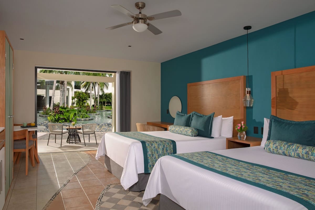 A hotel room with two beds, a ceiling fan, a view of a pool and palm trees, and a dining area with a table and chairs.