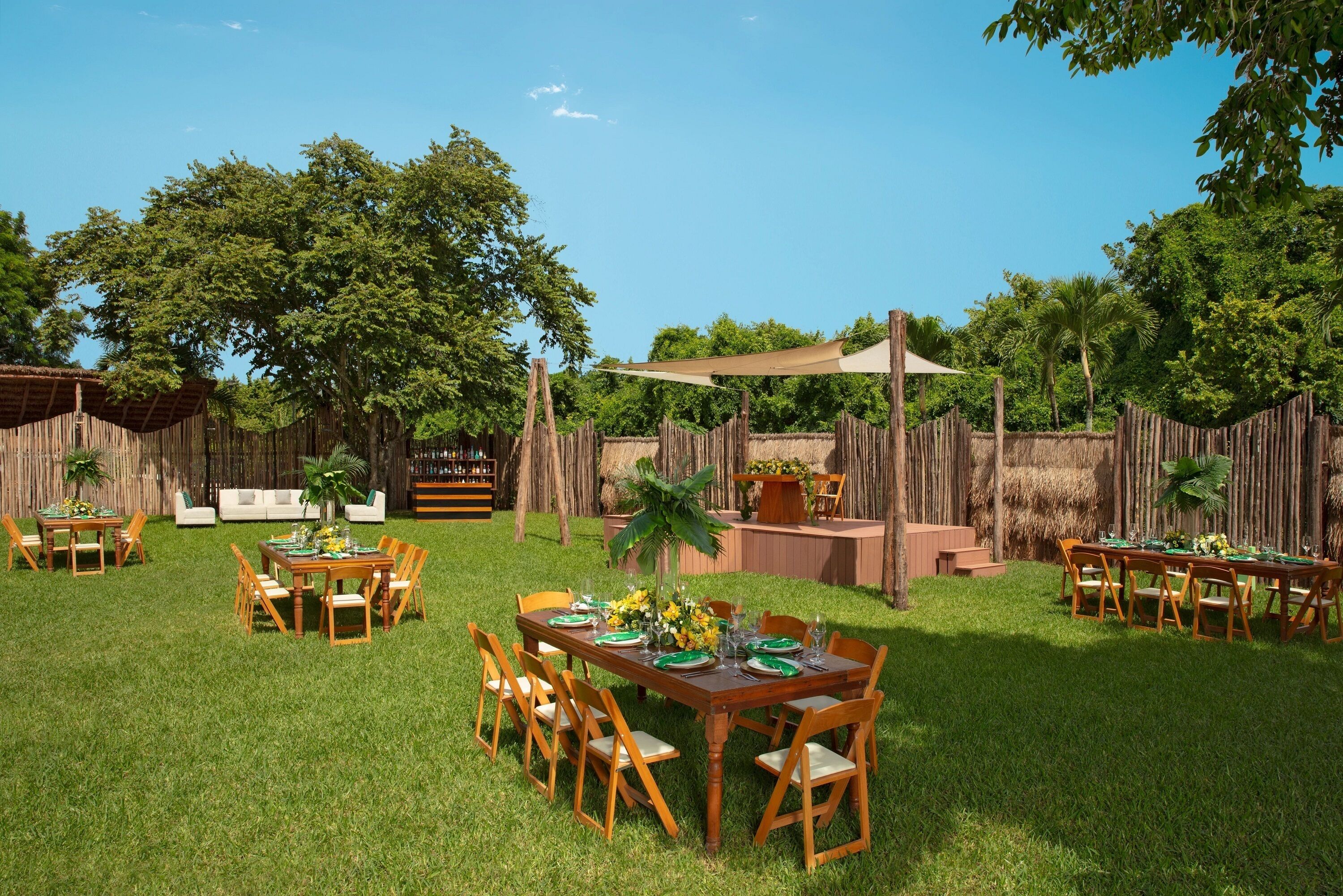 An outdoor dining area with wooden tables and chairs, surrounded by a wooden fence and lush greenery.