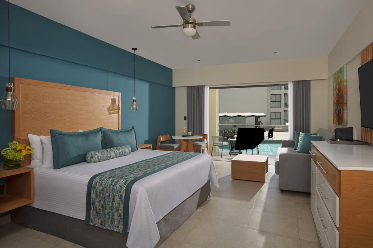 A modern hotel room with a large bed, a desk, a dining area, and a view of the city.