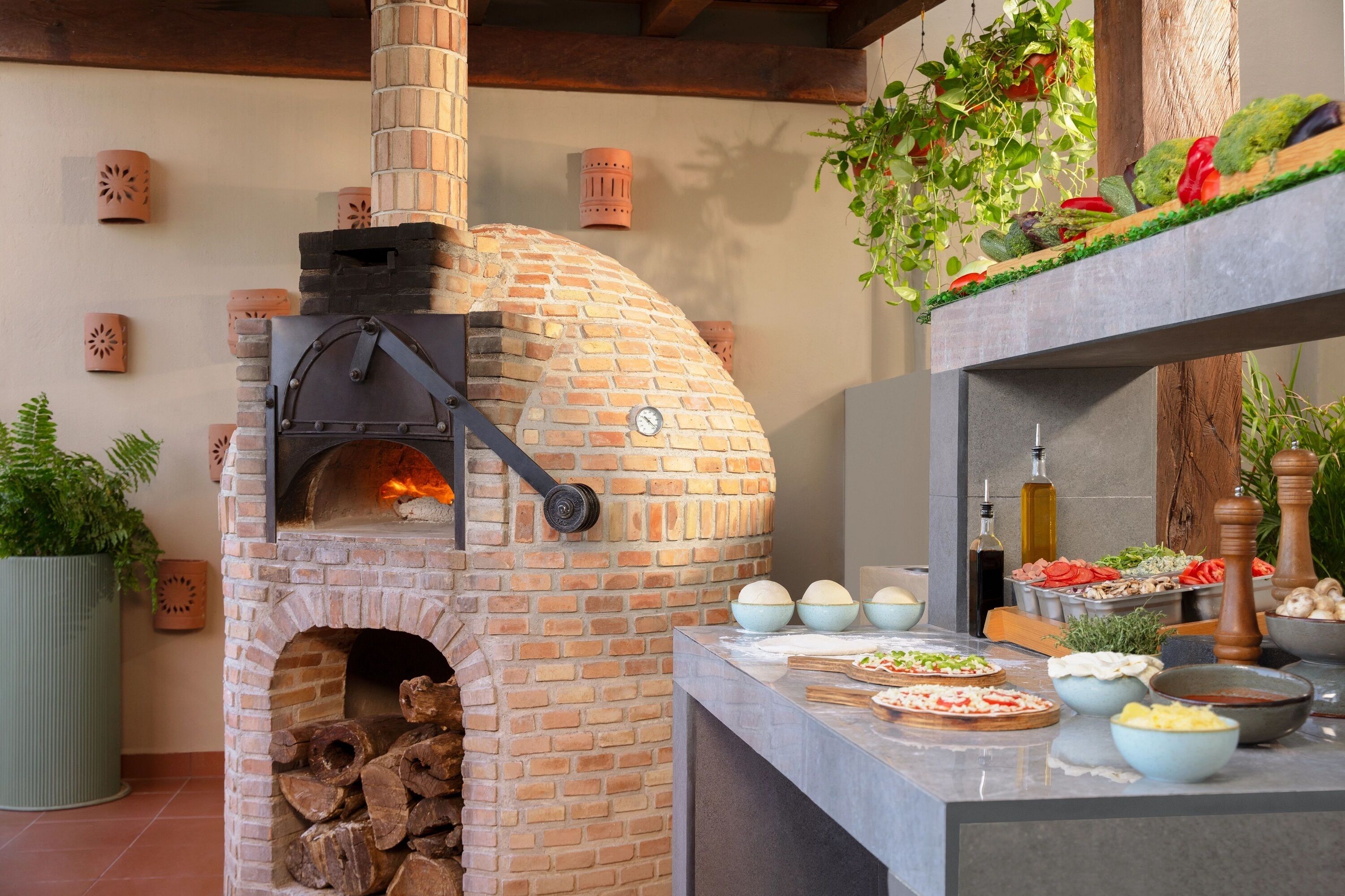 A brick oven pizza oven with a timer, surrounded by various bowls of ingredients and a wooden shelf with vegetables.