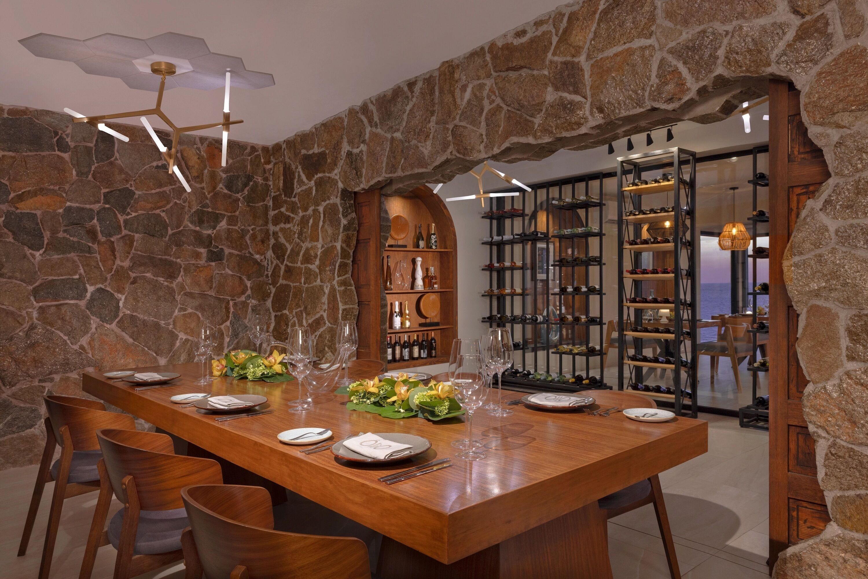 A dining area with a stone wall, wooden table, and chairs. There is a wine rack in the background.