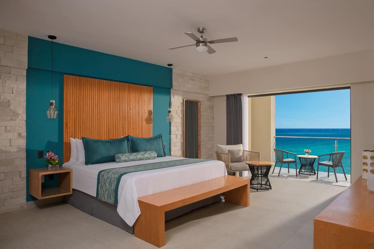 A bedroom with a large bed, a wooden headboard, a bench, a small table, and a view of the ocean.