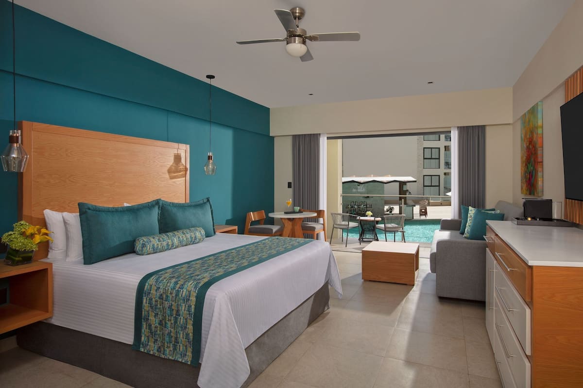 A modern hotel room with a large bed, a desk, a dining area, and a view of the pool.
