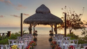 Un sendero de madera que conduce a una construcción de techo de paja con sillas blancas y arreglos florales.