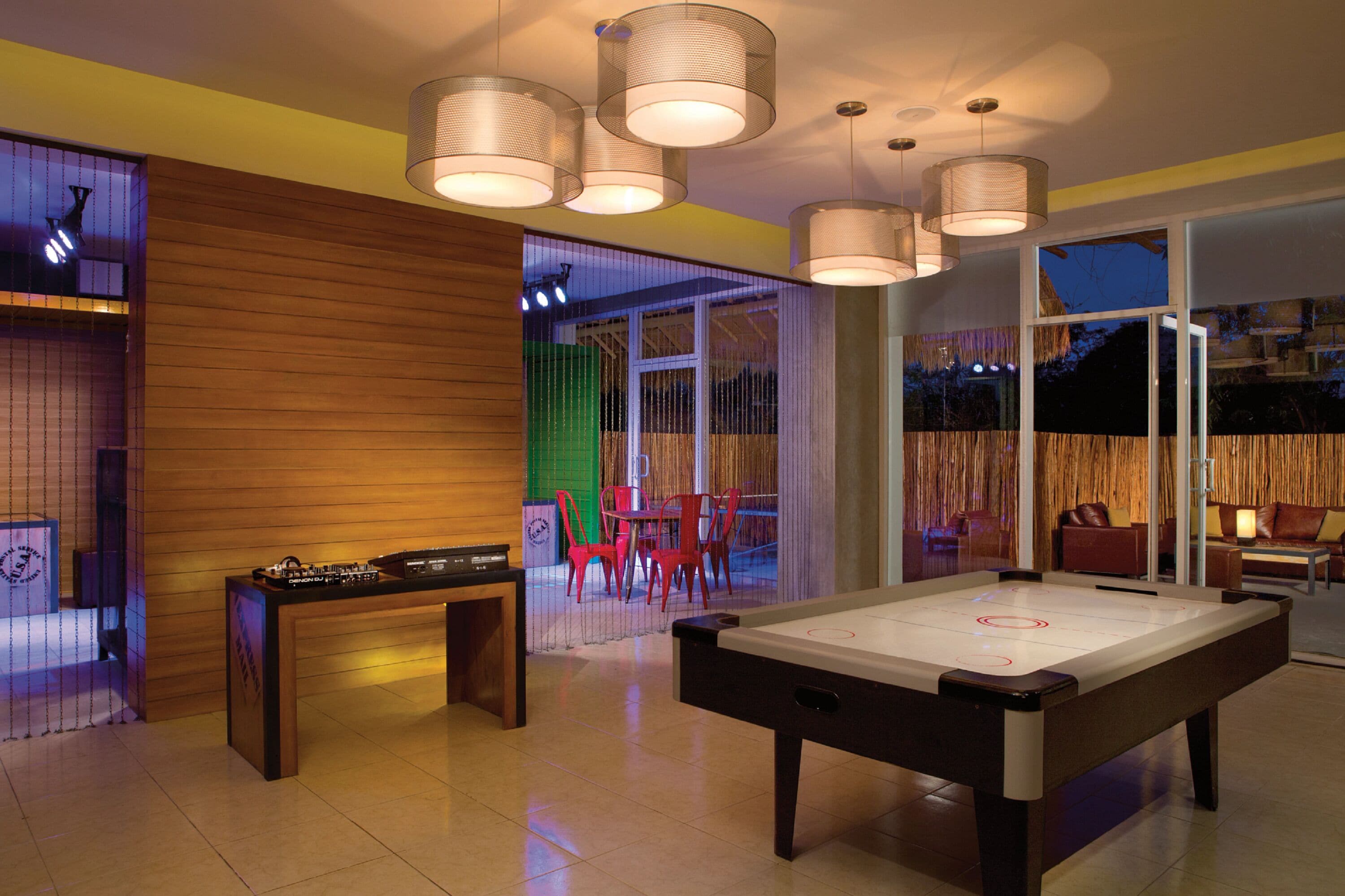 A pool table in a room with wooden paneling, a DJ setup, and a view of the outdoors through large windows.