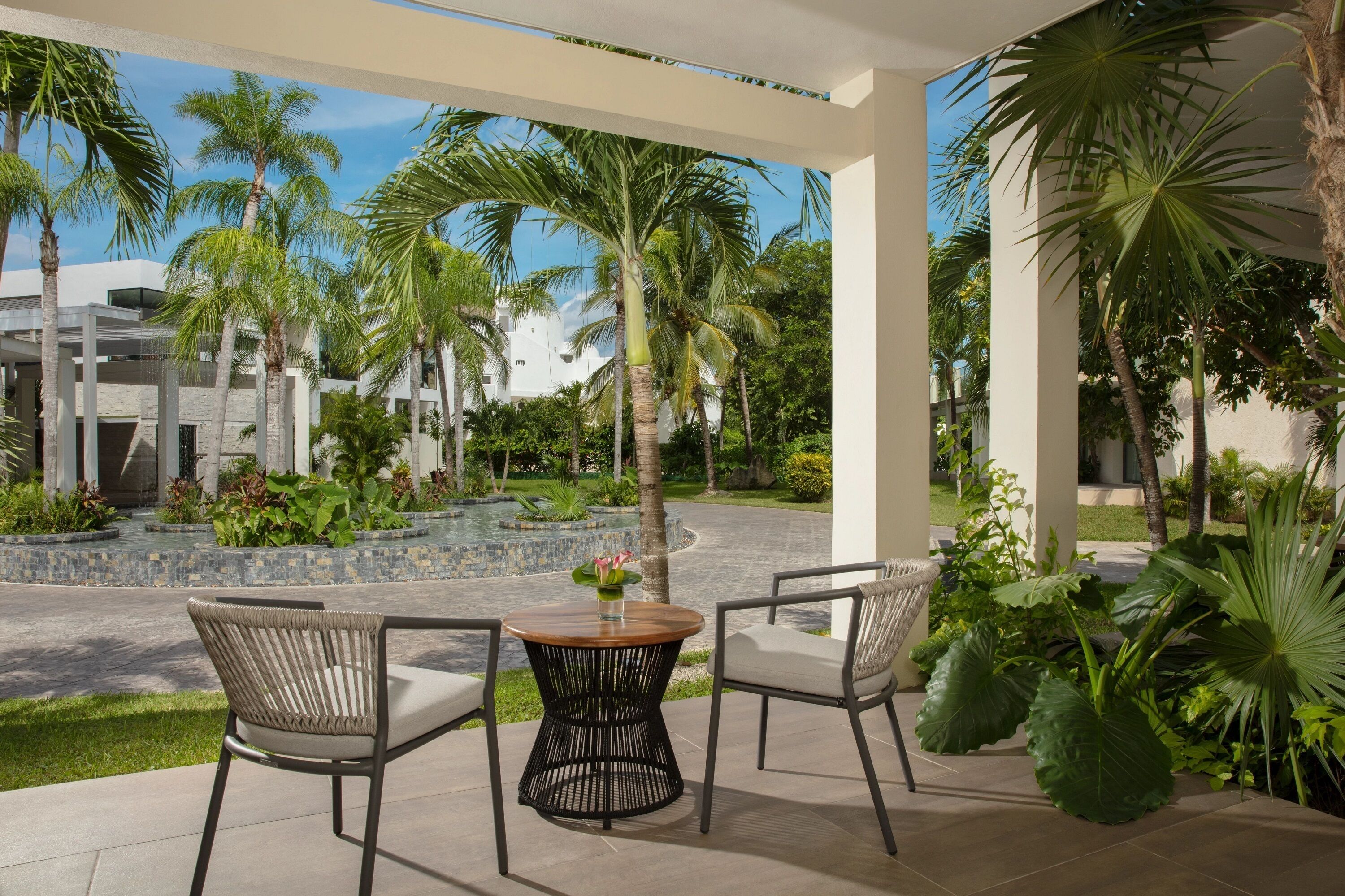 A covered outdoor seating area with two wicker chairs and a round table, surrounded by lush greenery and palm trees.