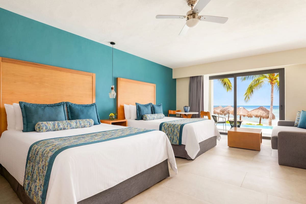 A hotel room with two beds, a ceiling fan, a view of the beach, and a balcony with a table and chairs.