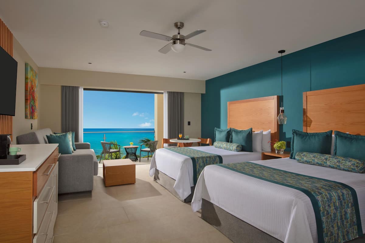 A hotel room with two beds, a balcony with a view of the ocean, a ceiling fan, and a small dining table.