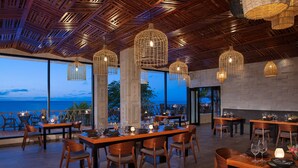 A restaurant with wooden tables and chairs, hanging wicker light fixtures, and large windows overlooking the ocean. - Dreams Cozumel Cape Resort & Spa - All Inclusive (Cozumel)