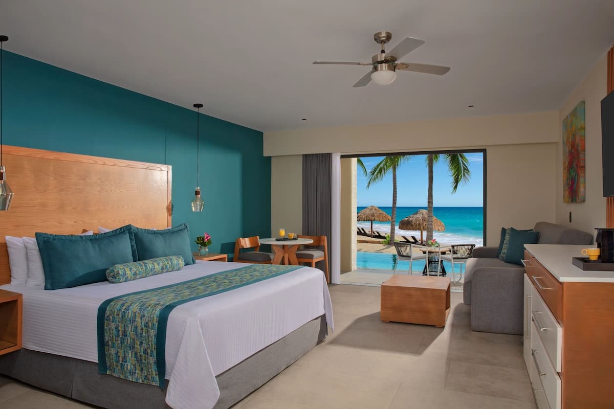 A modern hotel room with a large bed, a dining area, and a view of the beach.