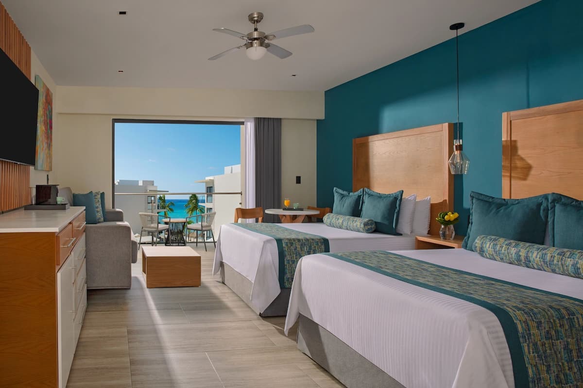 A hotel room with two beds, a kitchenette, a dining area, and a large window with a view of the ocean.
