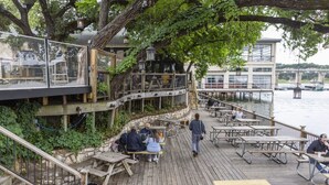 Um terraço de madeira com mesas de piquenique dando para um corpo d'água, ligado a um edifício com fachada de vidro.