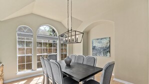 Uma sala de jantar com uma mesa de madeira escura, seis cadeiras cinzentas e um candelabro preto. Há grandes janelas com molduras brancas e vista para o verde lá fora.
