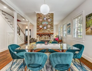 A dining area with a glass table, teal chairs, a brick fireplace, and a staircase.