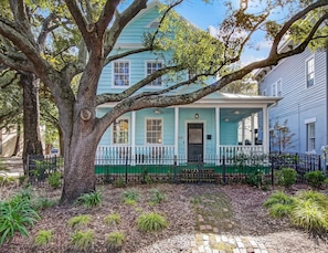 A light blue house with a porch, surrounded by trees and a brick pathway.