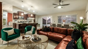 A modern living room with a leather sofa, green armchairs, and a coffee table. A kitchen with dark wooden cabinets and stainless steel appliances is visible in the background. - 4BR w pool, game room, playground (Phoenix)