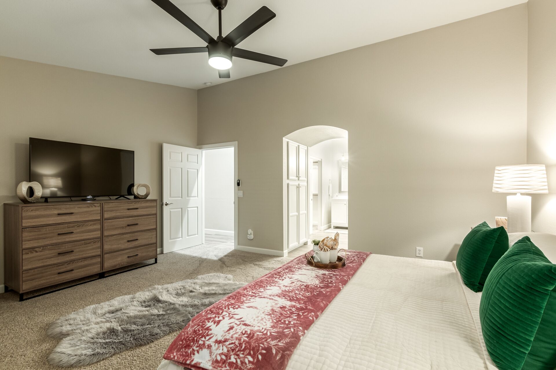 Une chambre à coucher comprenant un lit, une commode, un téléviseur, un ventilateur de plafond et un couloir menant à une autre pièce.