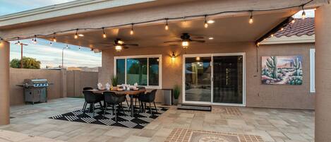 Un patio techado con una mesa y sillas, un ventilador de techo, luces colgantes y un cuadro en la pared.