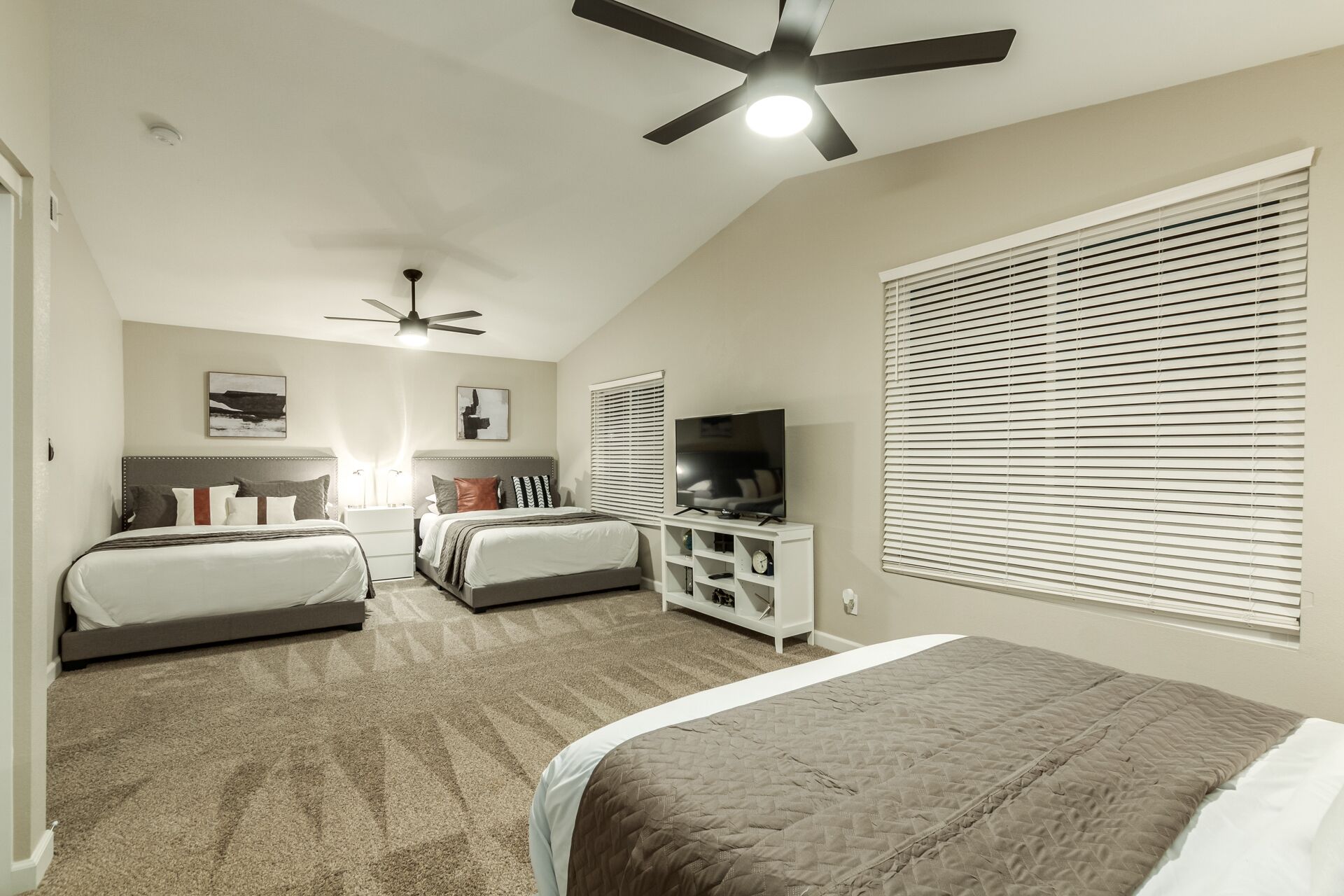 A bedroom with two beds, a ceiling fan, a TV, and a large window with blinds.