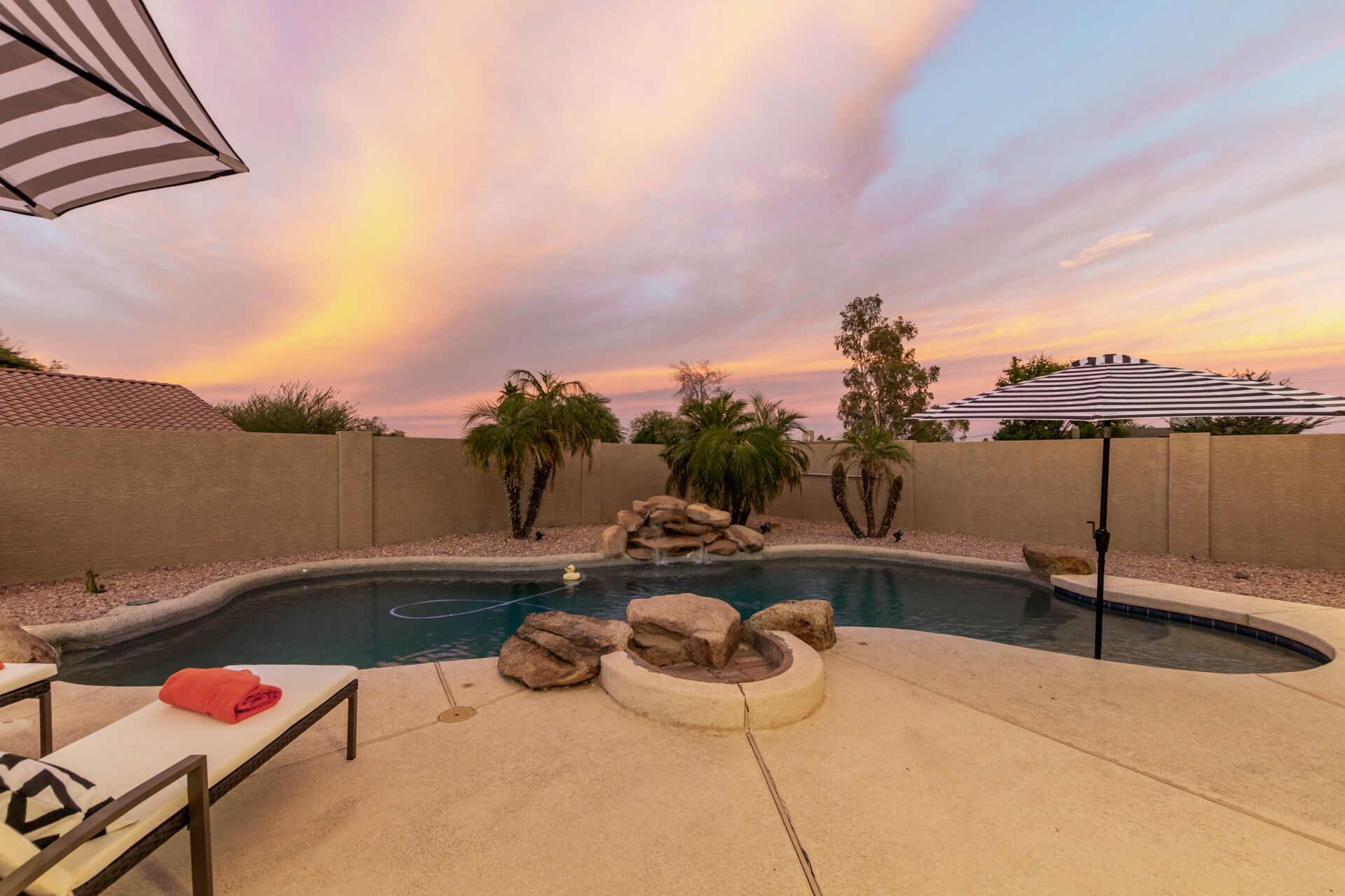 A swimming pool with a sunset view, surrounded by palm trees and a brick wall.