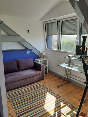 A compact living space with a sofa, a desk with a computer, and a window with blinds. - Seaside house (PRIMELIN)
