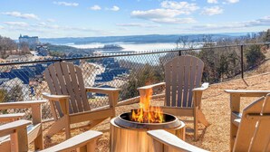 A deck with Adirondack chairs and a fire pit overlooking a scenic view of a lake and distant buildings. - Lakeview Retreat with Private Firepit, Pinball, Backyard, Hot Tub, Free Tickets & Steps to Amenities (Branson)
