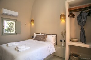 A bedroom with a bed, bedside lamps, a window, and a shelf with towels and a robe. - Pink Freud Villas Santorini (Santorini)