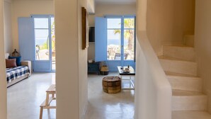 A spacious room with a staircase, a bed with a blue and white checkered bedspread, a wooden table, and a wicker ottoman. - Pink Freud Villas Santorini (Santorini)