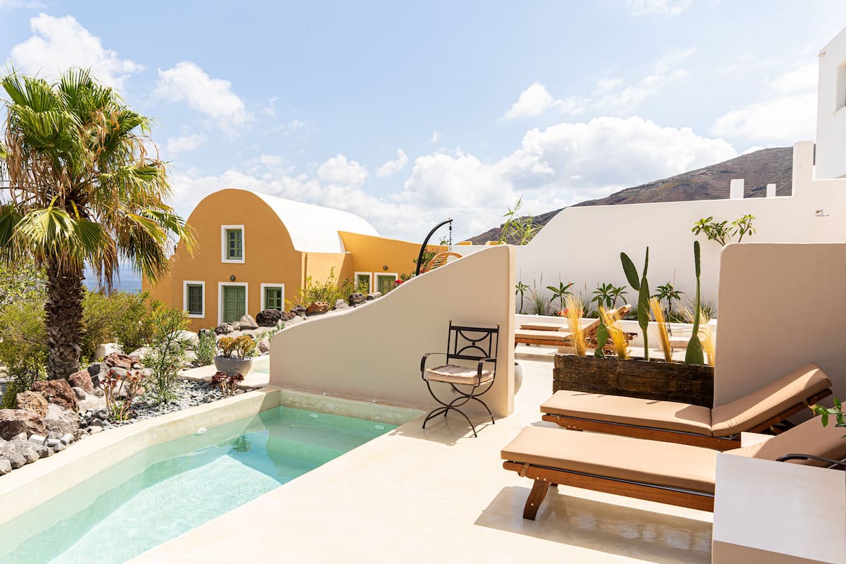 Zona junto a la piscina con sillas de descanso, una pequeña mesa y una silla. Hay plantas y una palmera cerca.