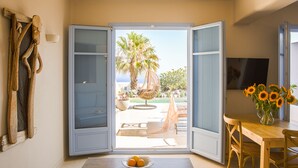 An open sliding door leading to a patio with a palm tree, a wicker chair, and a table with a bowl of fruit. - Pink Freud Villas Santorini (Santorini)