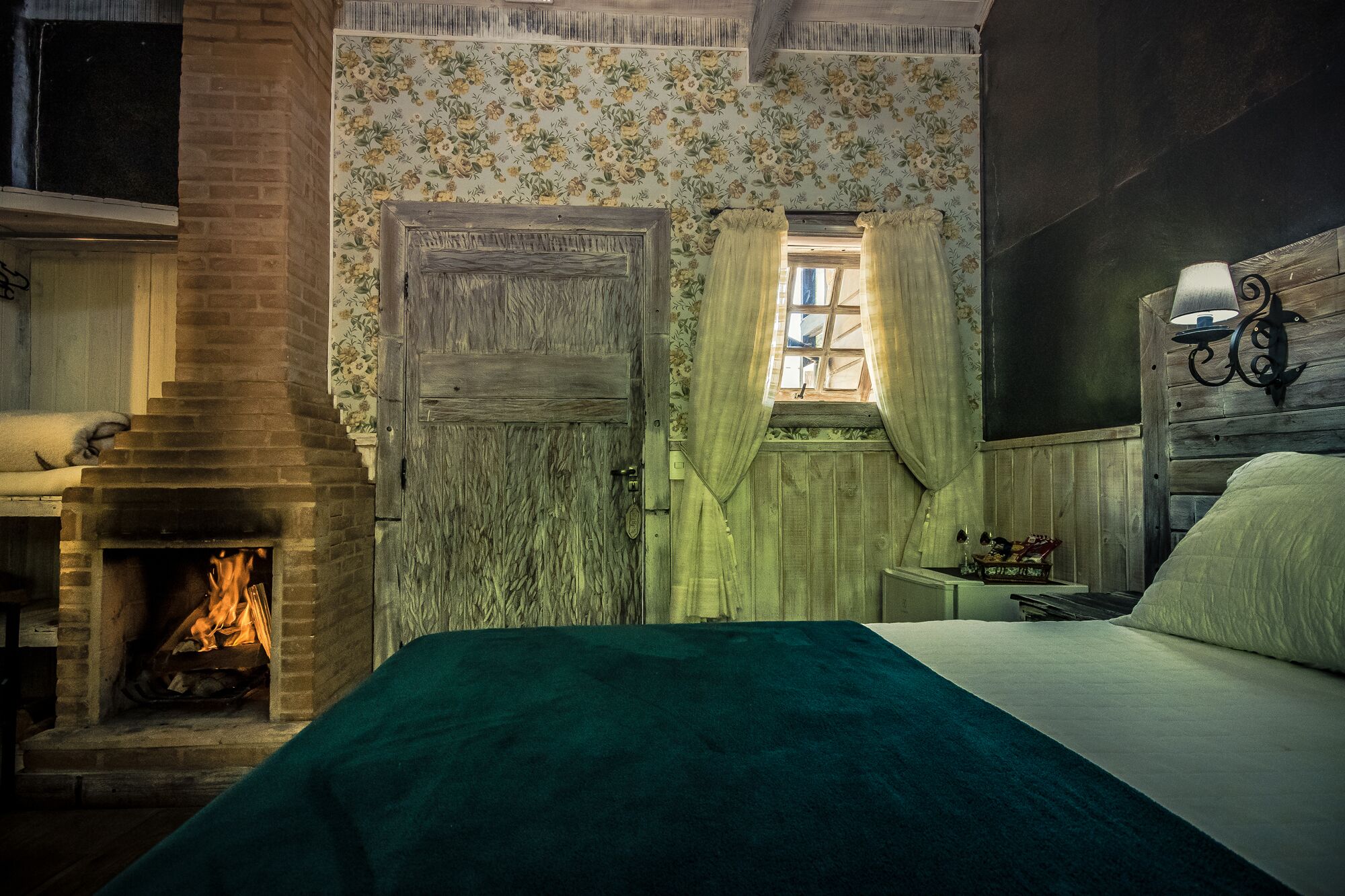 A cozy room with a fireplace, a bed, a window with curtains, and a wall-mounted lamp.