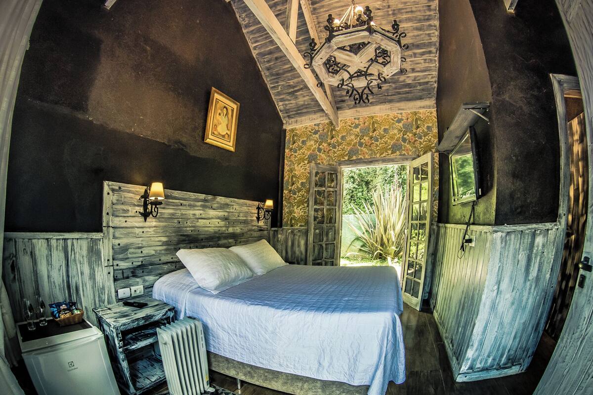 A rustic bedroom with a wooden headboard, a bed with a white bedspread, a radiator, a small table with a basket, and a window with a view of greenery.
