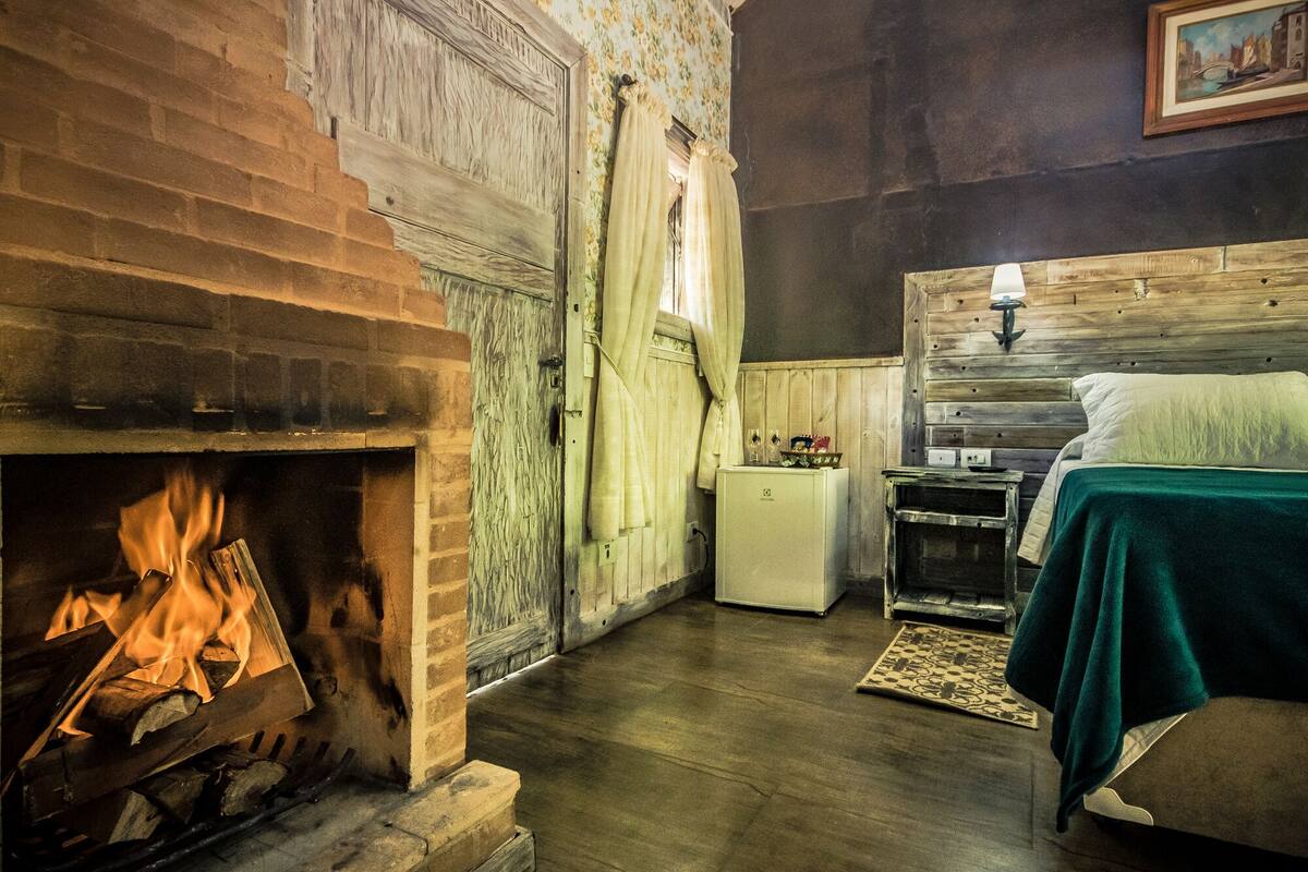 A rustic room with a fireplace, a bed, a small table, and a window with curtains.