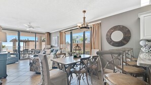 A spacious living area with a dining table, chairs, and a chandelier. There is a large window with a view of the outside. - Shoalwater 106 | Beachfront Corner Condo | Indoor, Outdoor Pools | My Beach Getaways (Orange Beach)