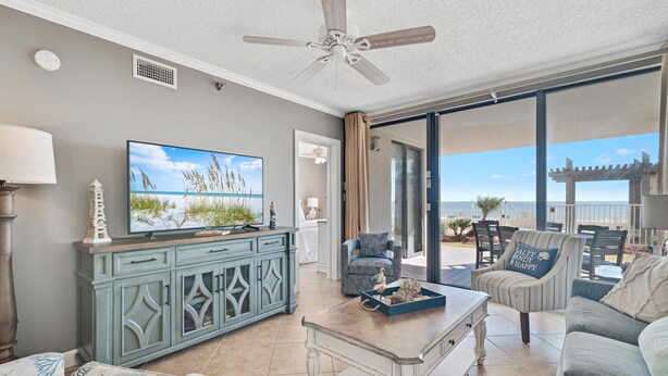 A living room with a television, a ceiling fan, and a view of the beach through large sliding glass doors. - Shoalwater 106 | Beachfront Corner Condo | Indoor, Outdoor Pools | My Beach Getaways (Orange Beach)