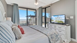 A bedroom with a large bed, a ceiling fan, a television on a dresser, and a view of the outdoors. - Shoalwater 106 | Beachfront Corner Condo | Indoor, Outdoor Pools | My Beach Getaways (Orange Beach)