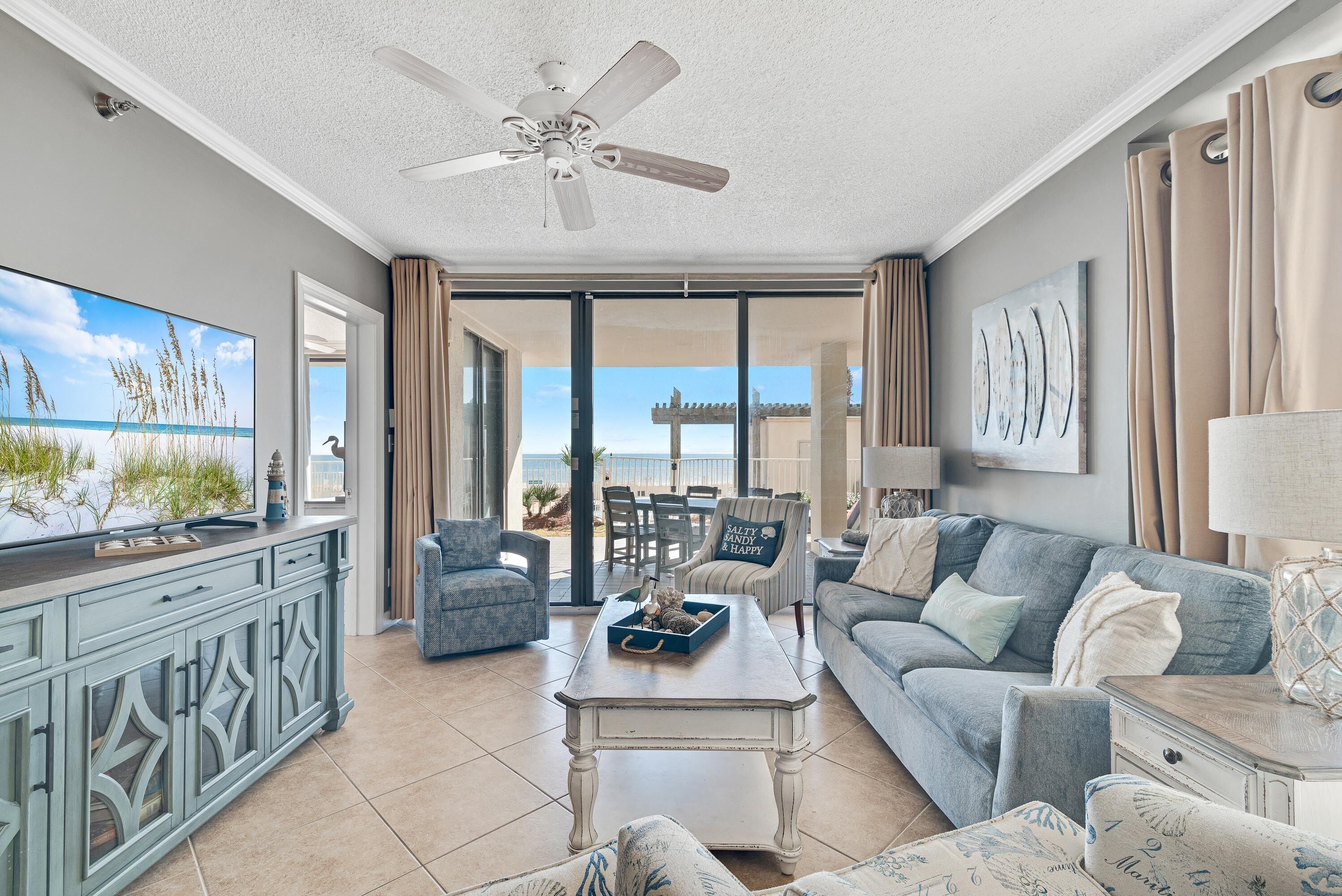 A spacious living room with a ceiling fan, a television, and a view of the beach through large sliding glass doors.