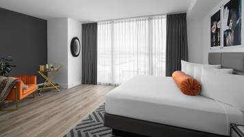 A modern hotel room with a large bed, a small orange armchair, a side table, and a large window with sheer curtains.